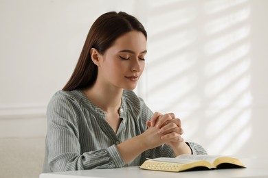 Photo of Religious young woman praying over Bible at table indoors