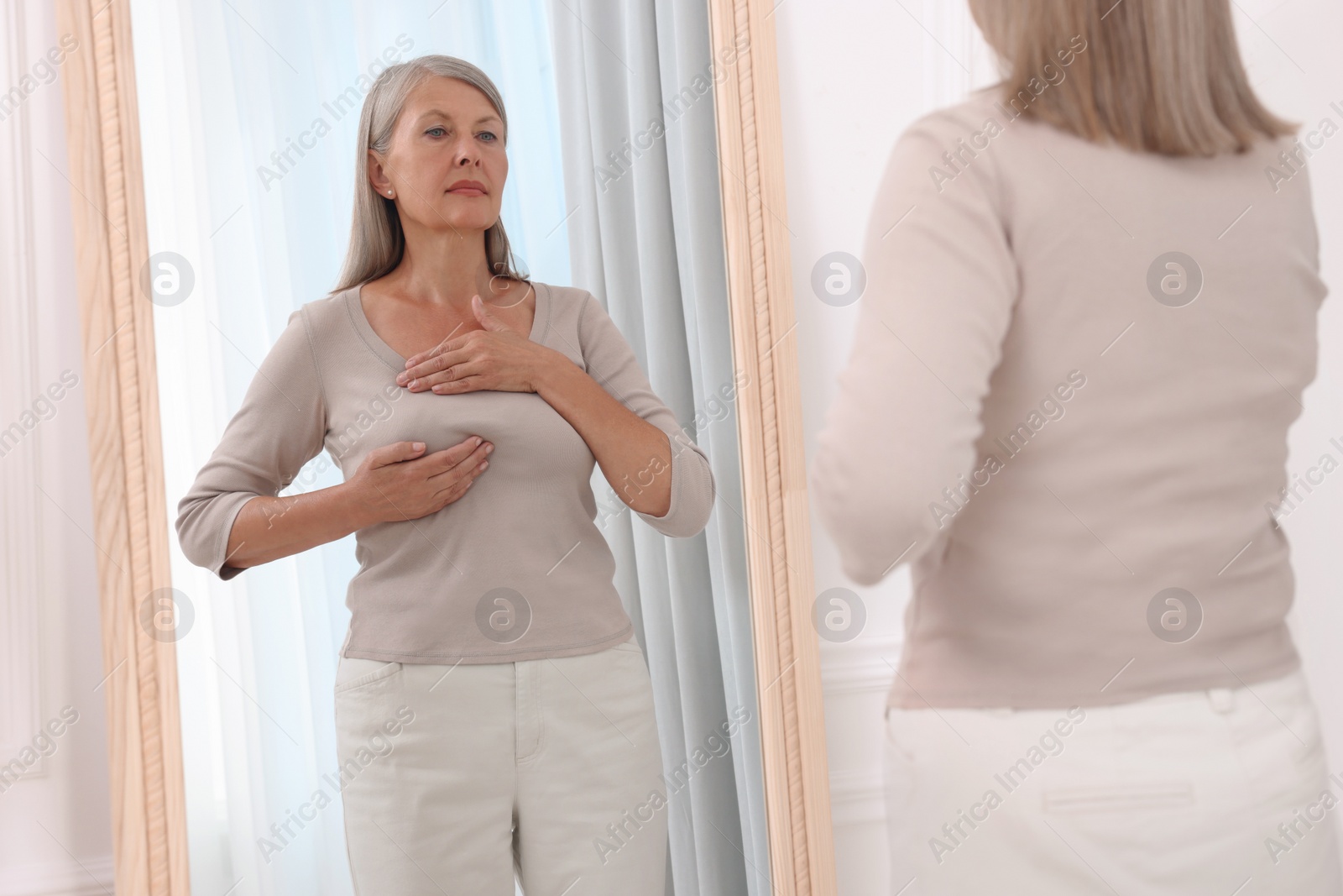 Photo of Beautiful senior woman doing breast self-examination near mirror indoors