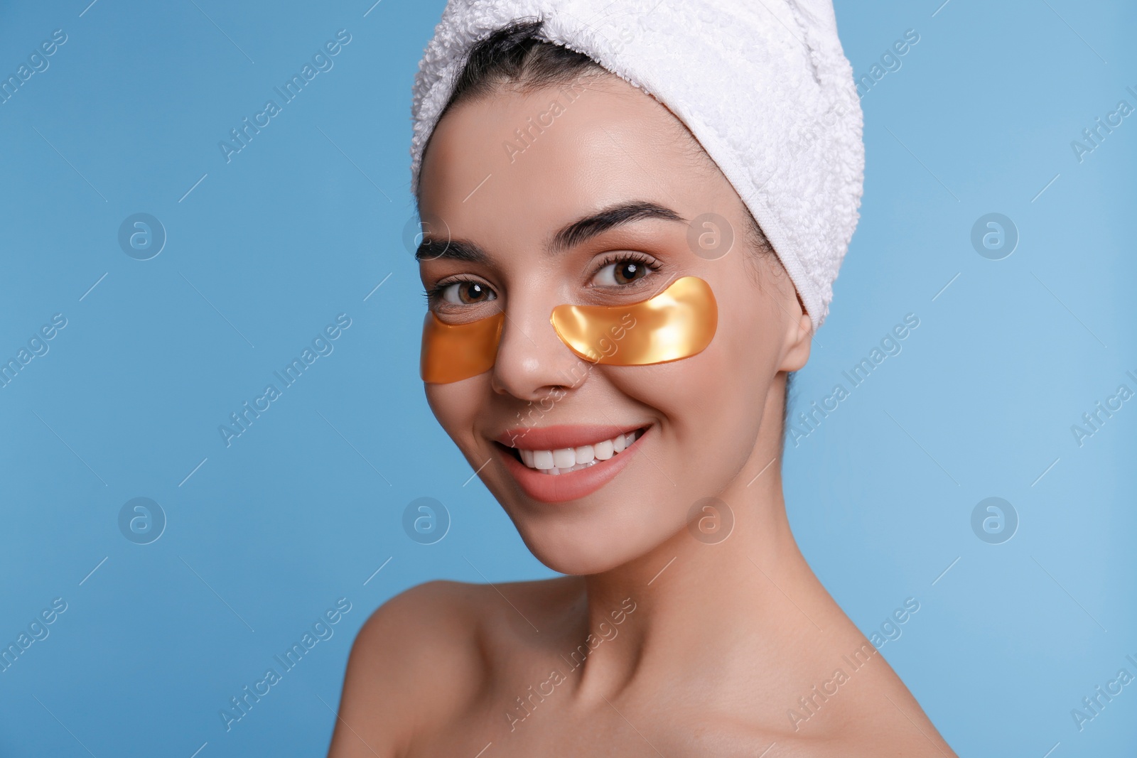 Photo of Beautiful young woman with under eye patches and hair wrapped in towel on light blue background