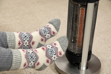 Photo of Woman warming legs near halogen heater indoors, closeup