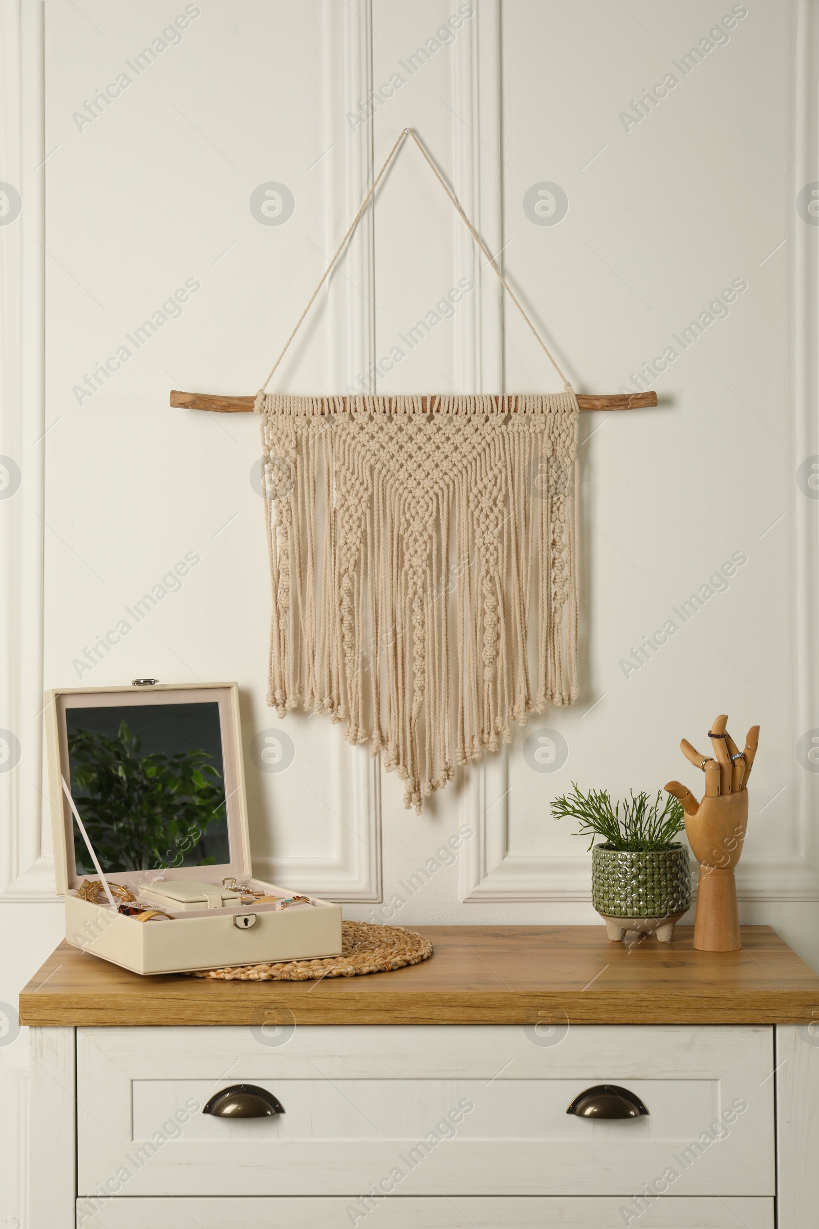 Photo of Open jewelry box and decor elements on chest of drawers near white wall with stylish macrame