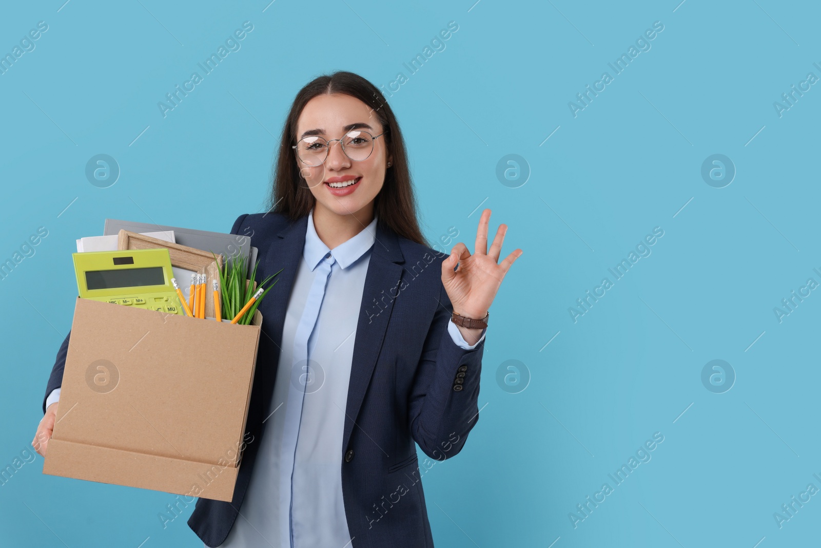 Photo of Happy unemployed woman with box of personal office belongings showing OK gesture on light blue background, space for text