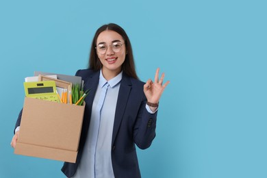 Photo of Happy unemployed woman with box of personal office belongings showing OK gesture on light blue background, space for text