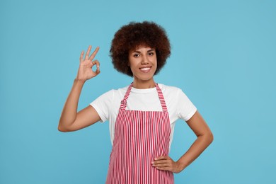 Photo of Happy young woman in apron showing ok gesture on light blue background