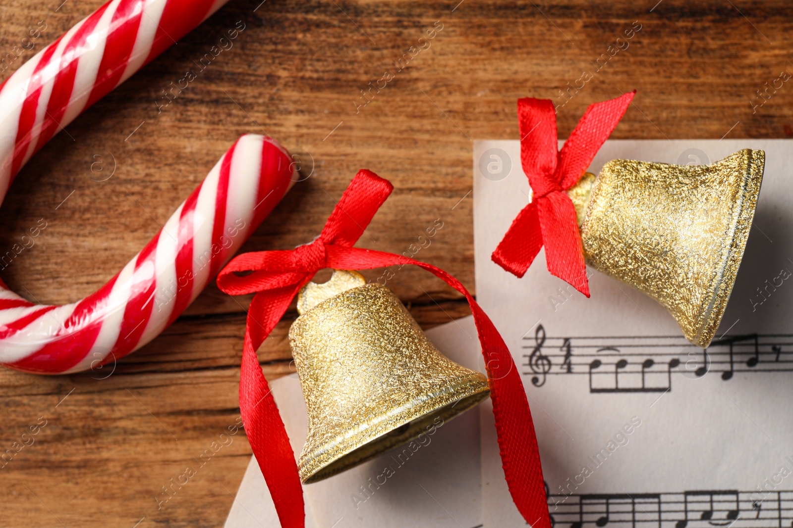 Photo of Golden shiny bells with red bows, music sheets and candy cane on wooden table, flat lay. Christmas decoration