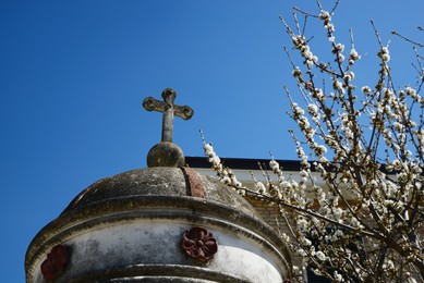Photo of Roof of old chapel with stone cross near blossoming tree against blue sky, closeup