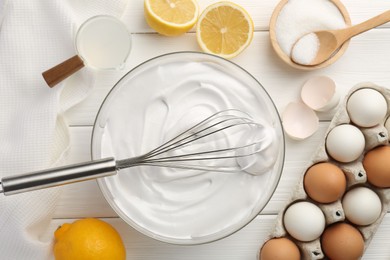 Bowl with whipped cream, whisk and ingredients on white wooden table, flat lay