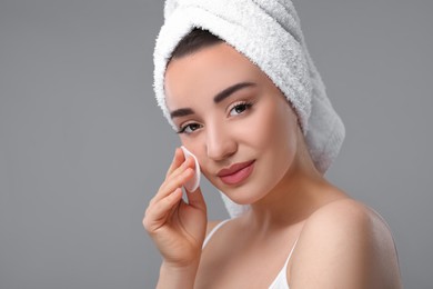 Photo of Beautiful woman in terry towel removing makeup with cotton pad on gray background, space for text