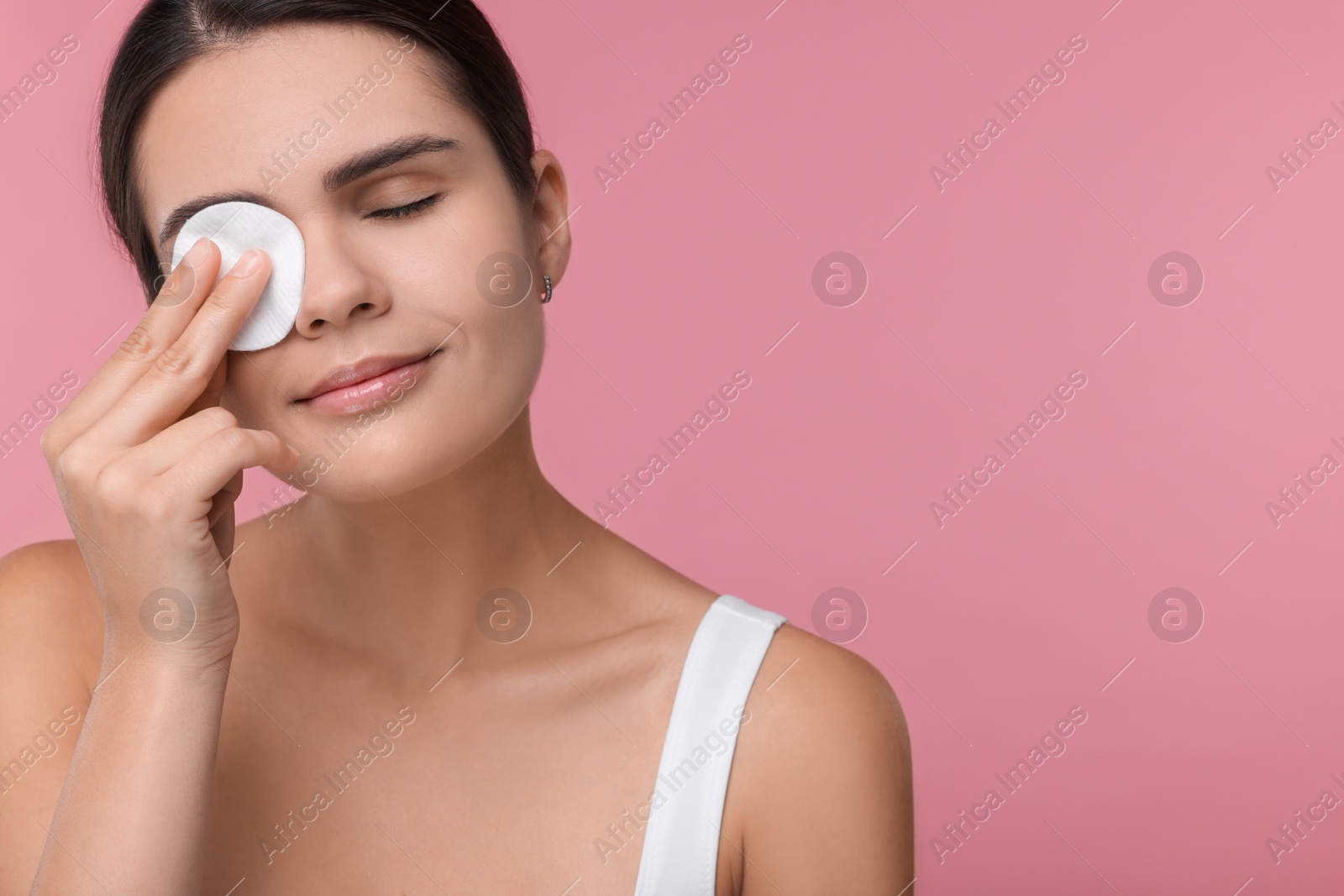 Photo of Young woman cleaning her face with cotton pad on pink background. Space for text