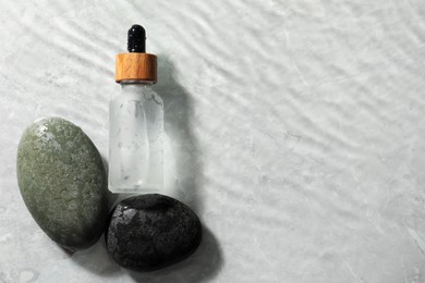 Photo of Bottle of face serum and spa stones in water on light background, flat lay. Space for text