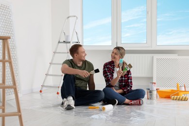 Photo of Happy couple with brushes and painting tools on floor in apartment during repair