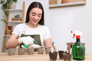Photo of Young woman spraying water onto vegetable seeds in peat pots at wooden table indoors