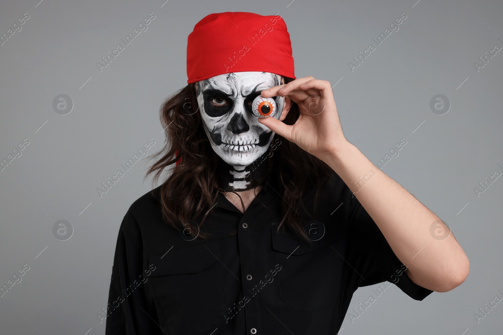 Photo of Man in scary pirate costume with skull makeup and decorative eyeball on light grey background. Halloween celebration