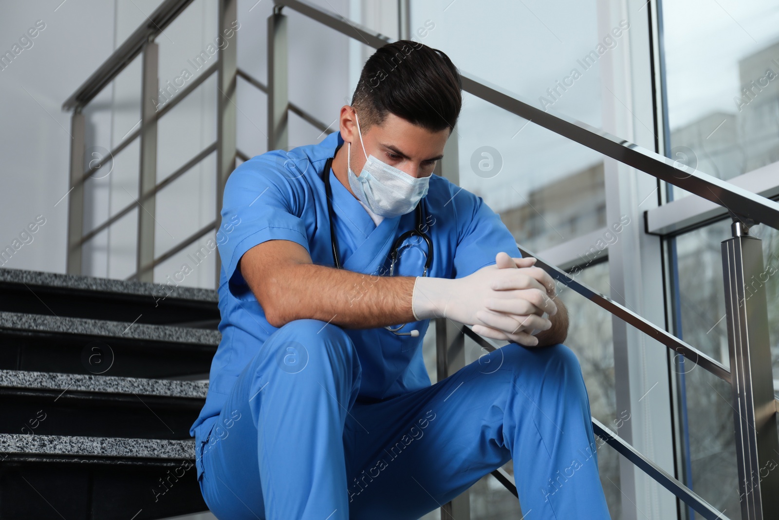 Photo of Sad doctor in facial mask on stairs indoors. Stress of health care workers during coronavirus pandemic