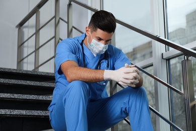 Photo of Sad doctor in facial mask on stairs indoors. Stress of health care workers during coronavirus pandemic