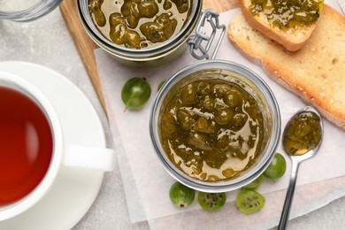 Jars with delicious gooseberry jam, fresh berries, toast and tea on grey table, flat lay