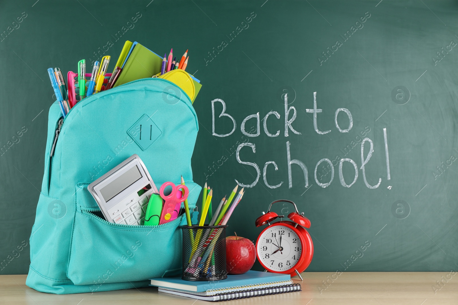 Photo of Bright backpack with school stationery on wooden table near green chalkboard. Back to School