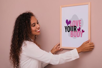 Image of Stop body shaming and love yourself. Smiling woman hanging body positive picture on beige wall indoors