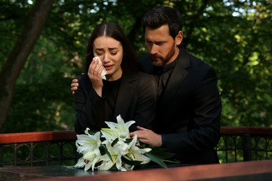 Photo of Sad couple mourning near granite tombstone with white lilies at cemetery. Funeral ceremony