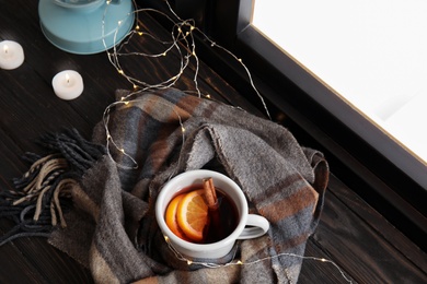 Photo of Composition with cup of hot winter drink, scarf and Christmas lights on wooden sill near window. Cozy season
