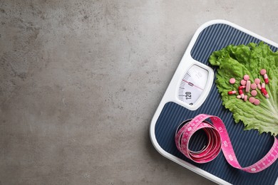 Photo of Scales with weight loss pills, lettuce leaf and measuring tape on grey table, top view. Space for text