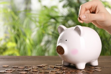 Woman putting coin into piggy bank at wooden table against blurred green background, closeup. Space for text