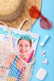 Photo of String bag with magazine, earphones and summer accessories on light blue background, flat lay