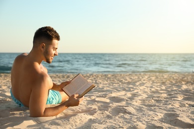 Young man reading book on sandy beach near sea