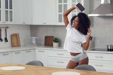 Photo of Beautiful woman with smartphone wearing stylish underwear and t-shirt dancing in kitchen, space for text