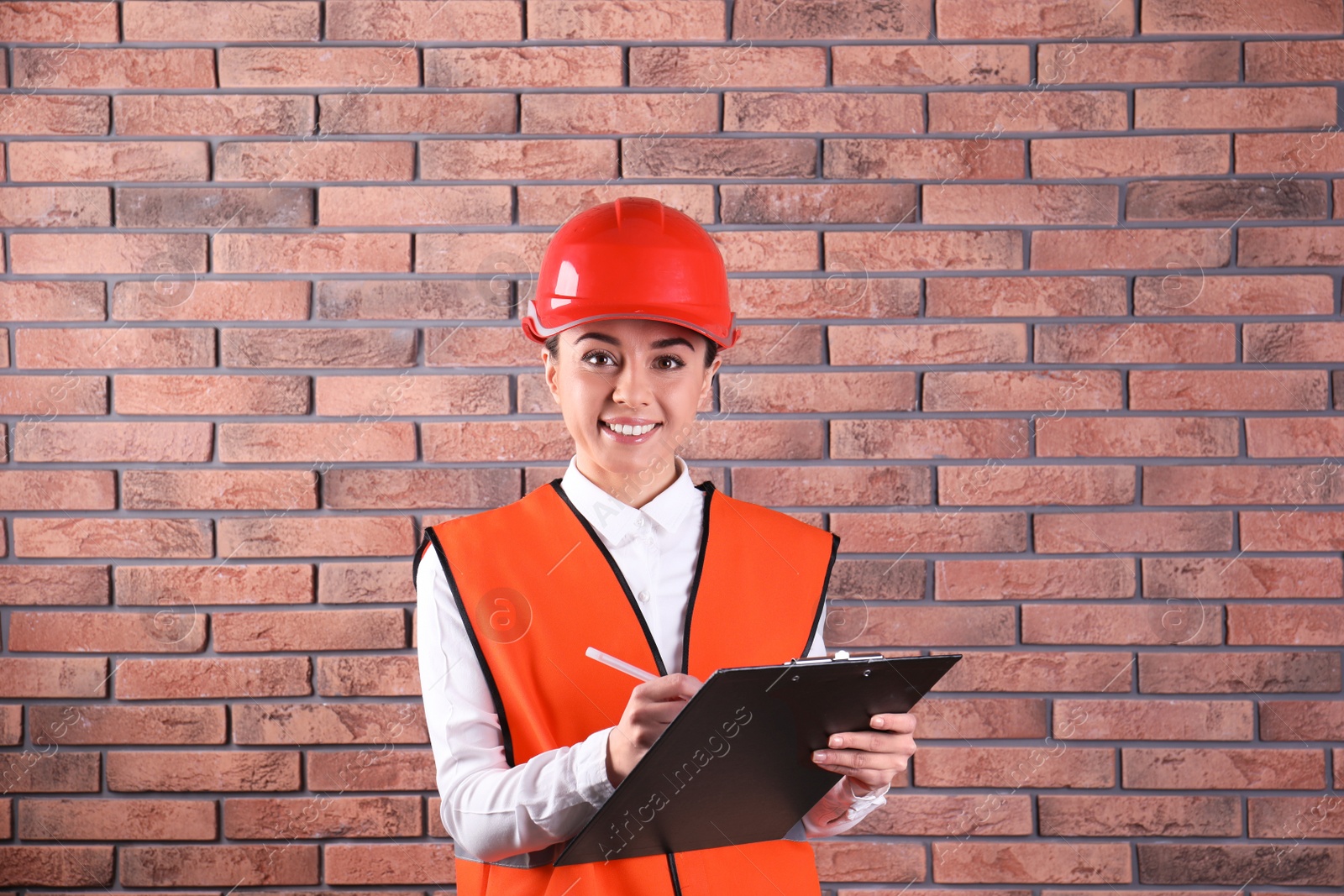 Photo of Female industrial engineer in uniform with clipboard on brick wall background. Safety equipment