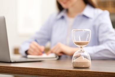 Hourglass with flowing sand on desk. Woman taking notes while using laptop indoors, selective focus