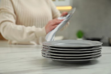 Woman wiping dish with towel at white marble table in kitchen, focus on stack of plates