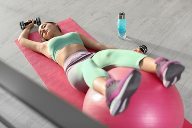 Lazy young woman with sport equipment on yoga mat indoors