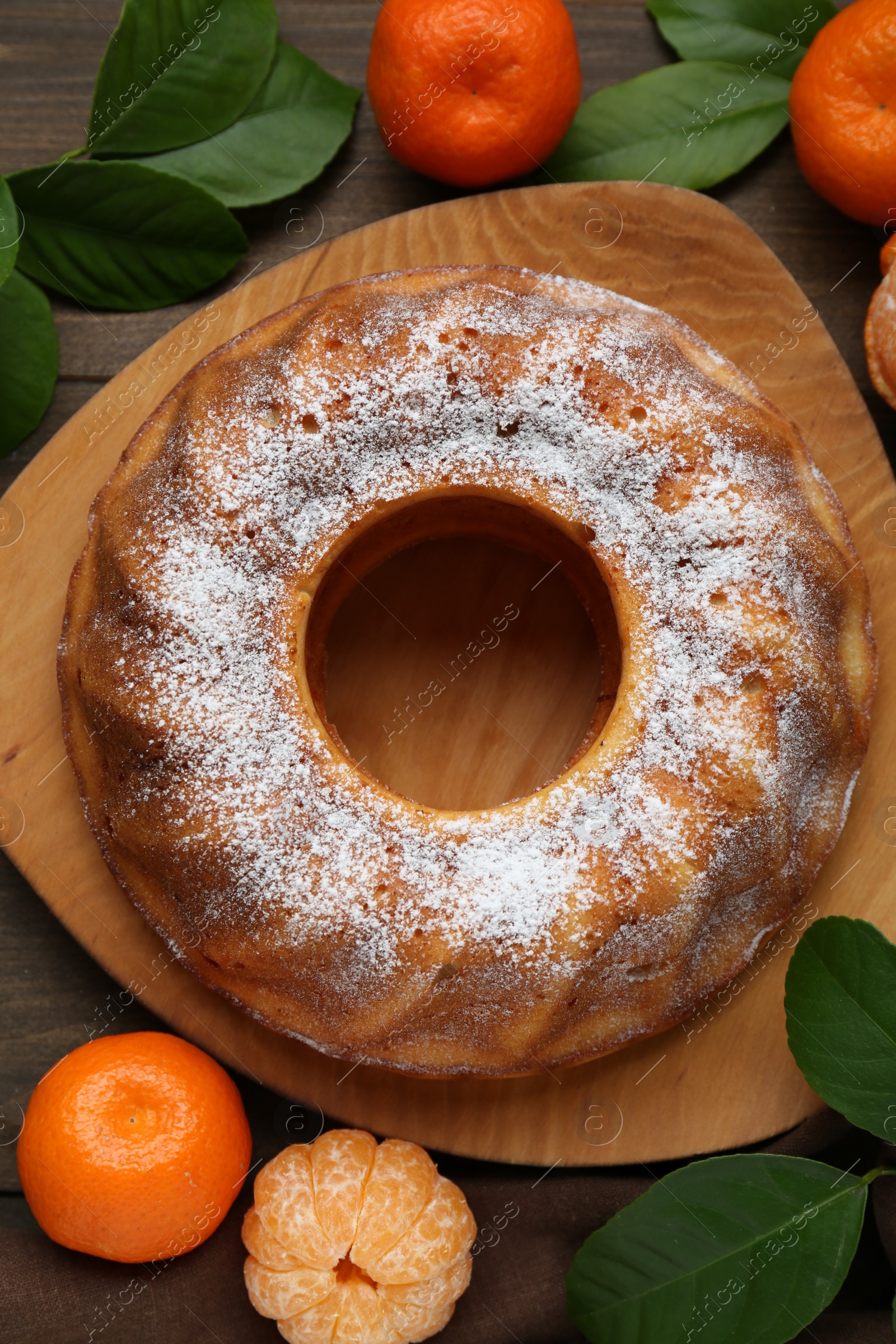 Photo of Homemade yogurt cake with tangerines, powdered sugar and green leaves on wooden table, flat lay