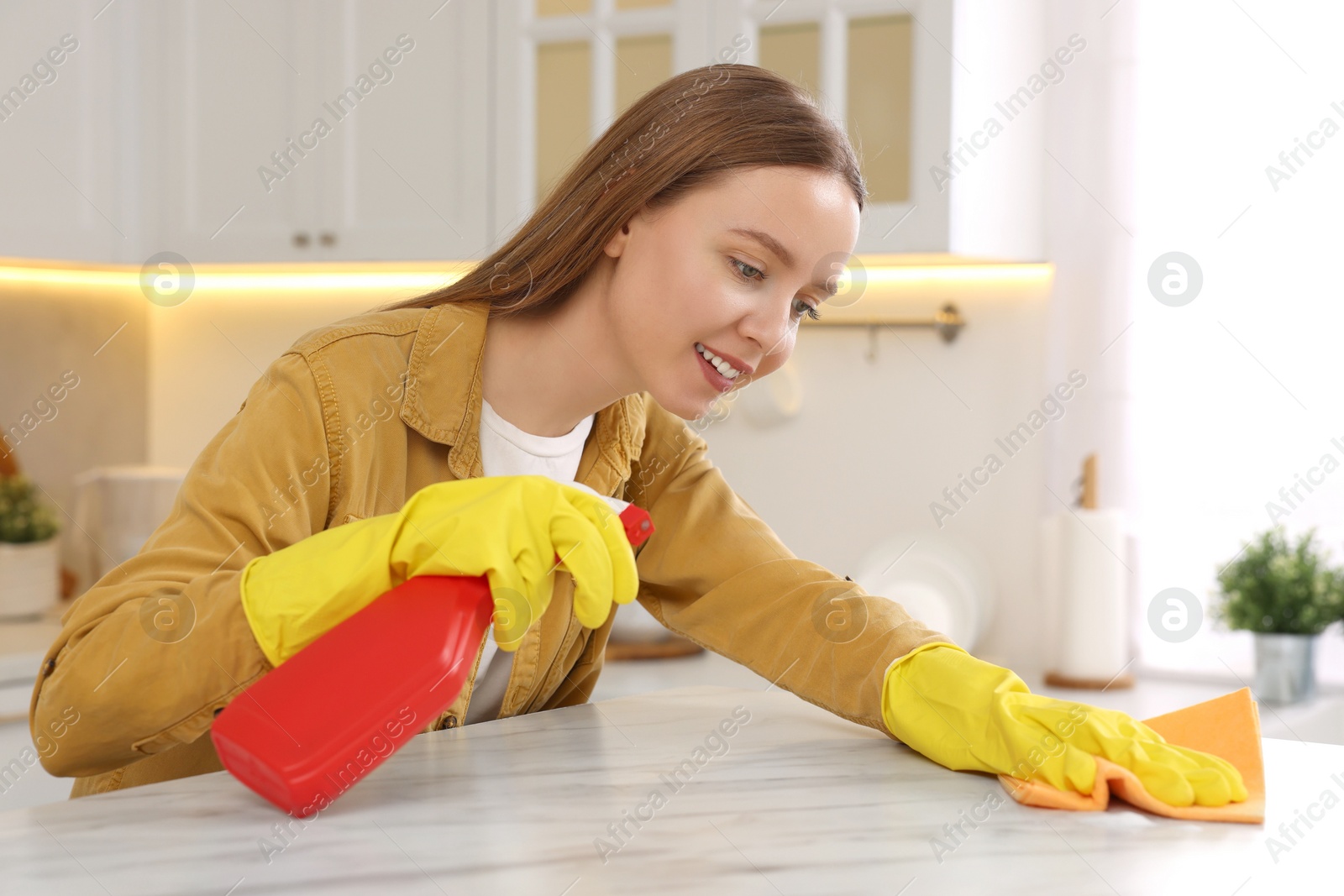 Photo of Woman with spray bottle and microfiber cloth cleaning white marble table in kitchen