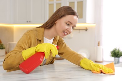Woman with spray bottle and microfiber cloth cleaning white marble table in kitchen