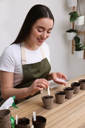 Photo of Woman inserting cards with names of vegetable seeds into peat pots at table indoors