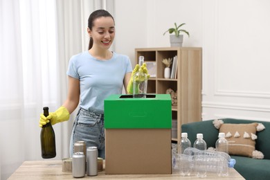 Photo of Garbage sorting. Smiling woman throwing glass bottle into cardboard box in room