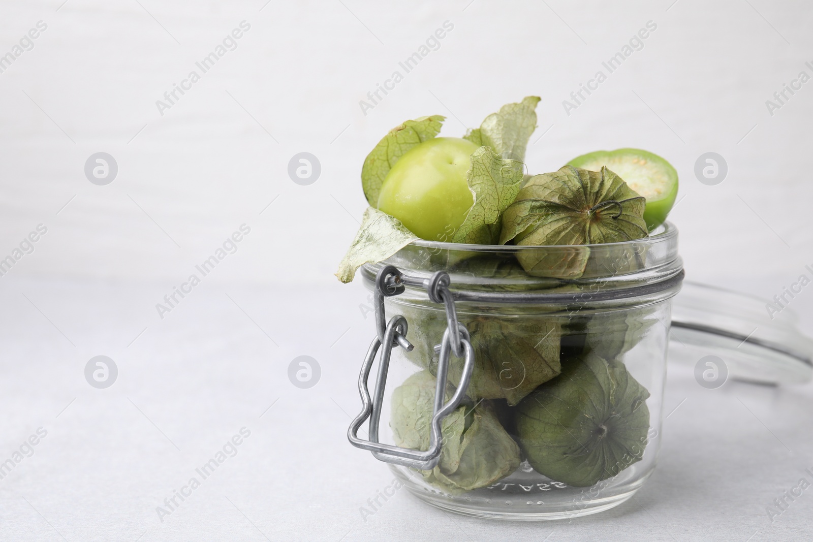 Photo of Fresh green tomatillos with husk in glass jar on light table, space for text