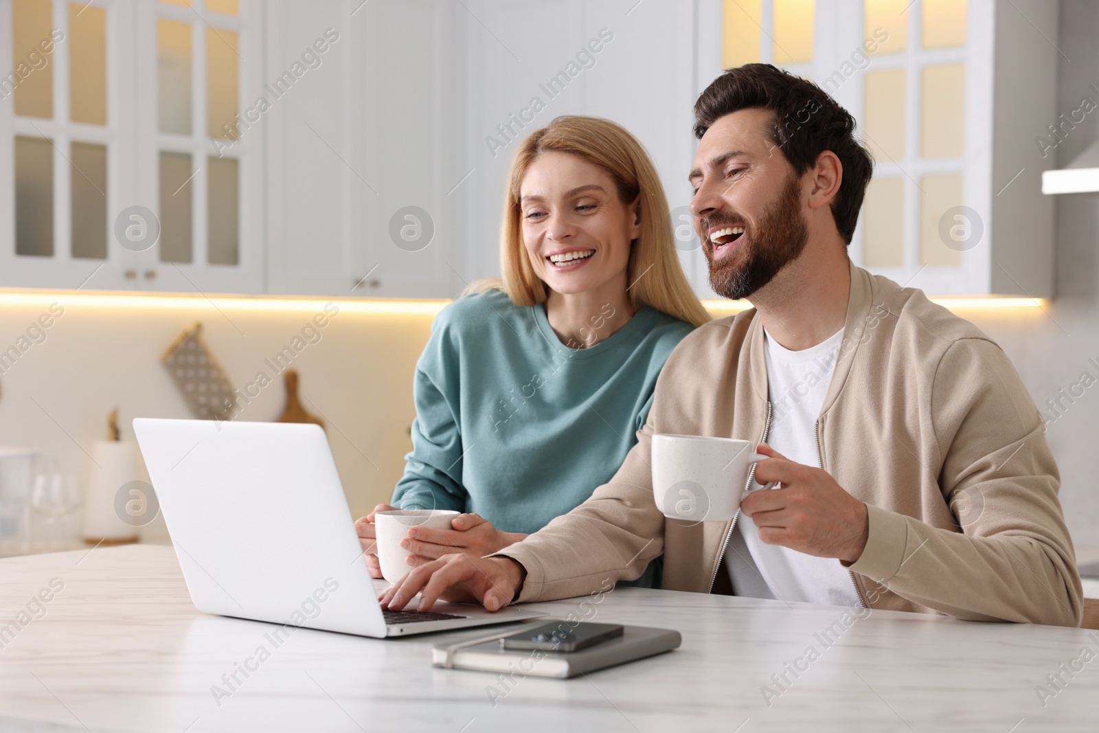 Photo of Happy couple with laptop and cups of drink at white table in kitchen