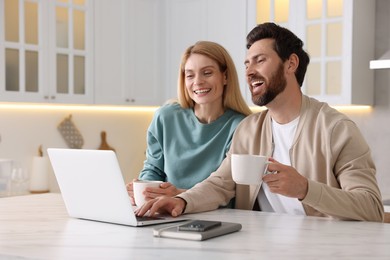 Happy couple with laptop and cups of drink at white table in kitchen