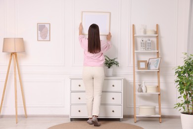 Photo of Woman hanging picture frame on white wall at home, back view