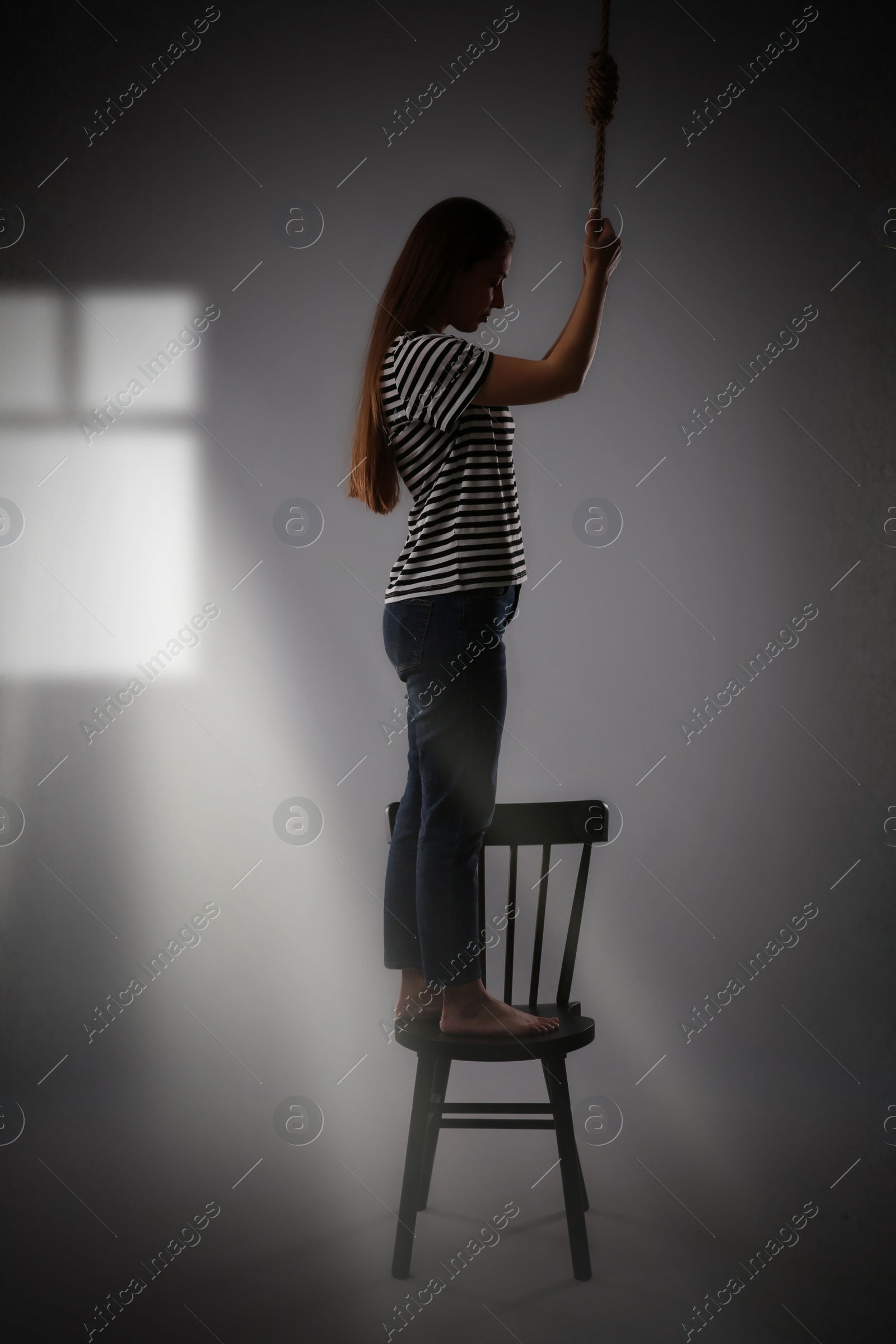 Image of Depressed woman with rope noose standing on chair in room