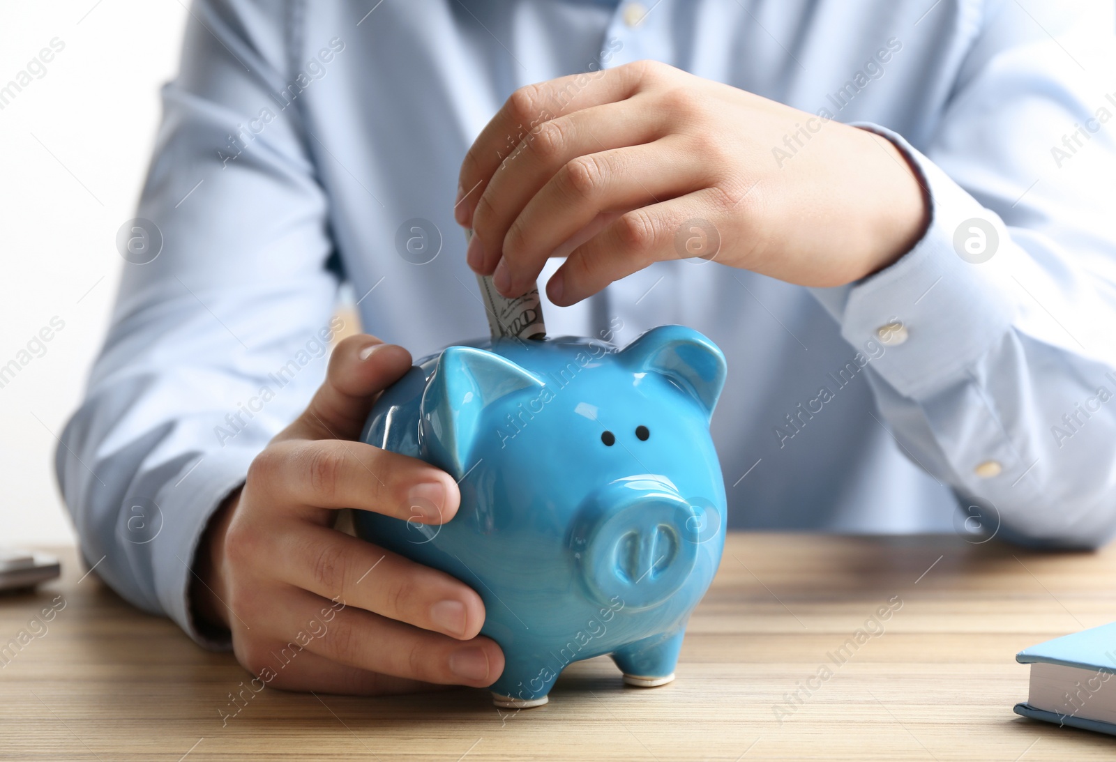 Photo of Man putting dollar banknote into piggy bank at wooden table indoors, closeup. Money savings