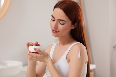 Photo of Beautiful young woman applying body cream onto shoulder in bathroom