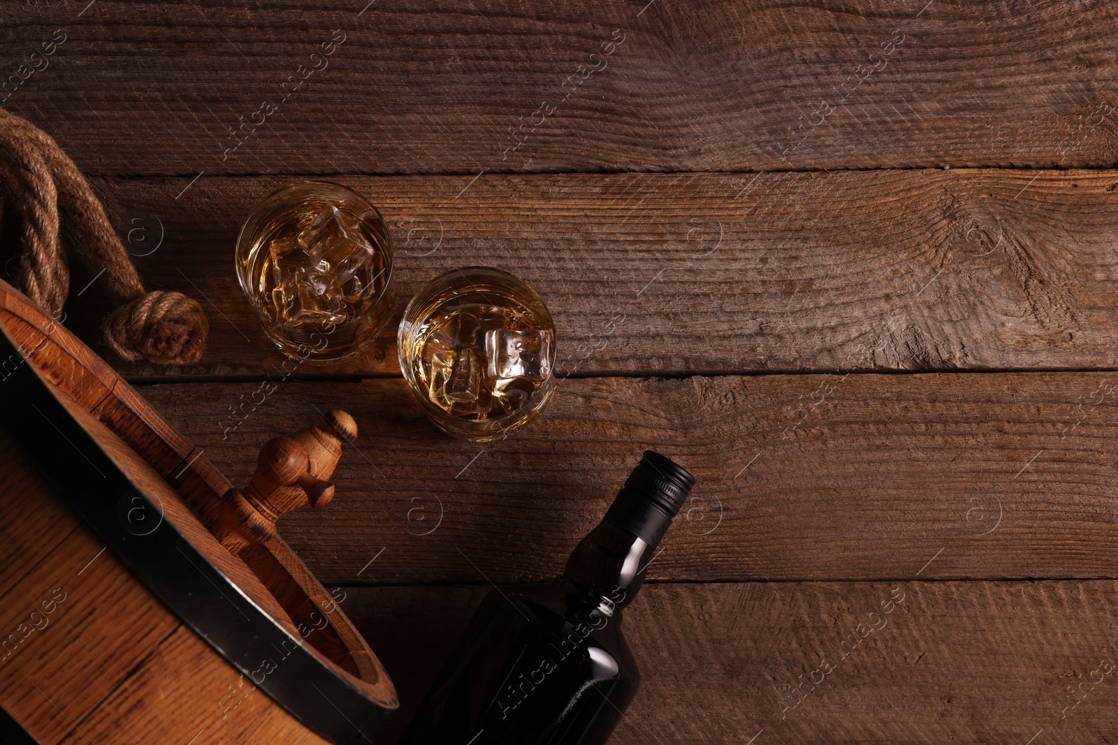 Photo of Whiskey with ice cubes in glasses, bottle and barrel on wooden table, flat lay. Space for text