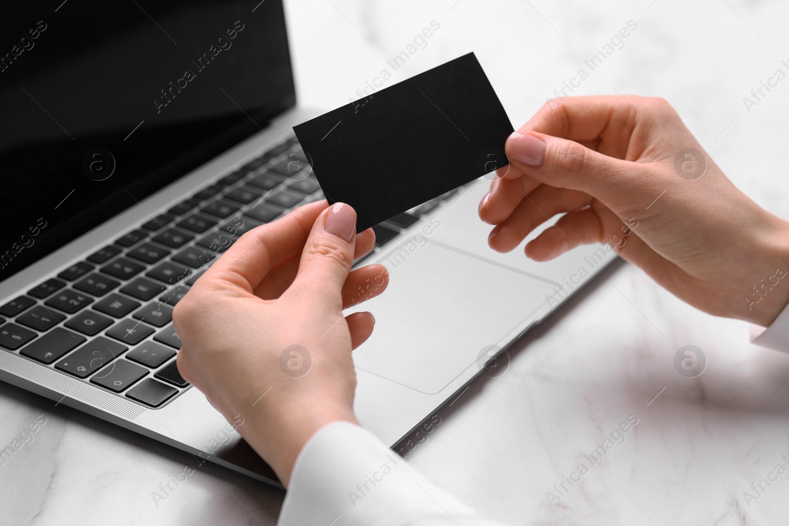 Photo of Woman with laptop holding blank business card at white table, closeup. Space for text