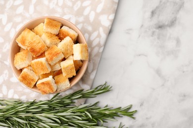 Photo of Delicious crispy croutons in bowl and rosemary on white marble table, flat lay. Space for text