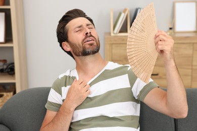 Bearded man waving hand fan to cool himself on sofa at home