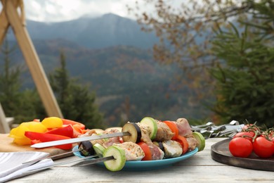 Metal skewers with raw marinated meat and vegetables on wooden table against mountain landscape